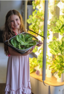 Young girl standing in front of a Gardyn Home System holding a basket of harvested greens.